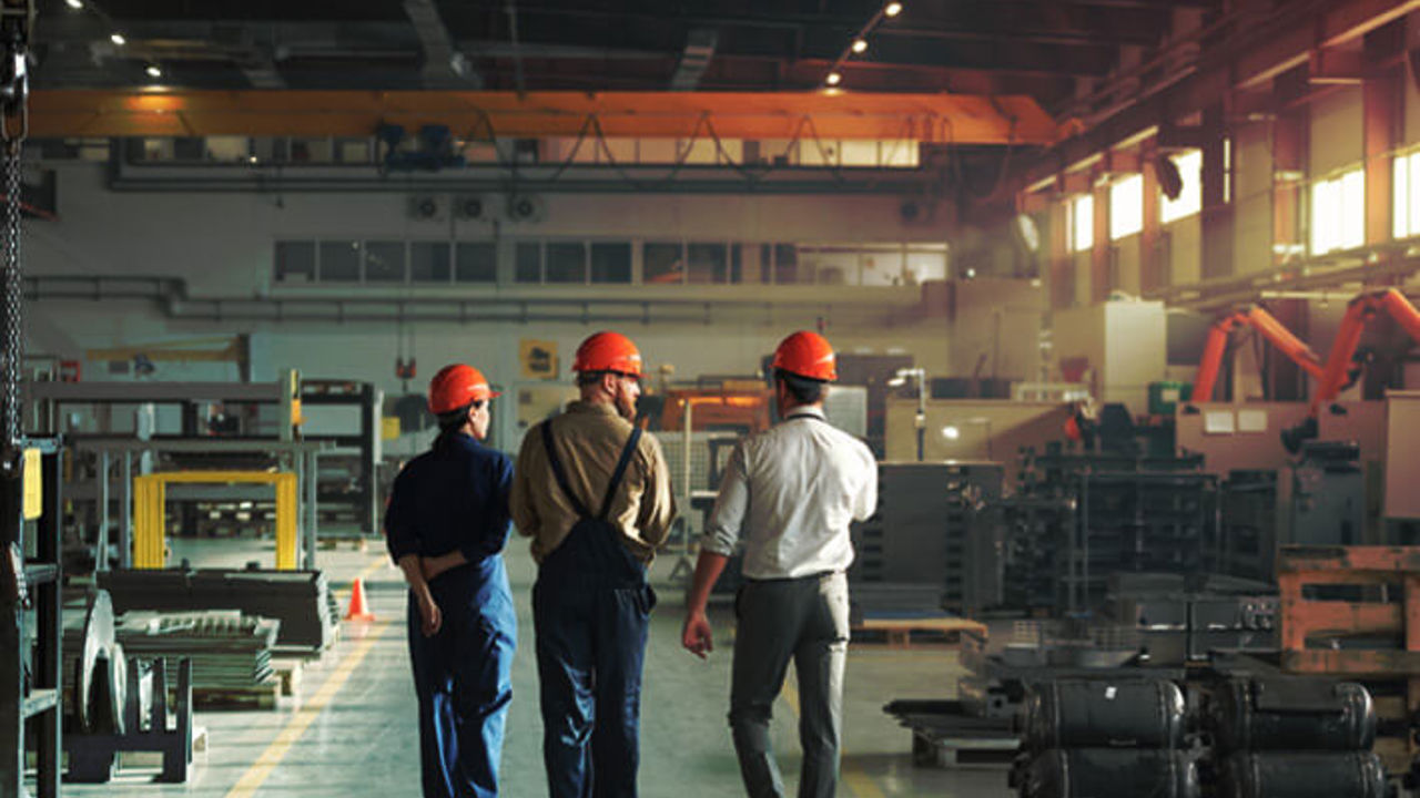 Three workers in helmets and overalls walk through a spacious, well-lit factory. Metal parts and equipment are organized on pallets and shelves. A large overhead crane is visible above, and a safety cone is placed near the path.