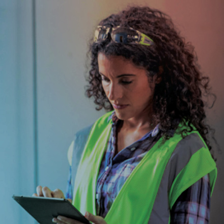 A person with curly hair wearing a neon safety vest and glasses on their head is using a tablet. They are standing against a blue-gray background, appearing focused on the device.