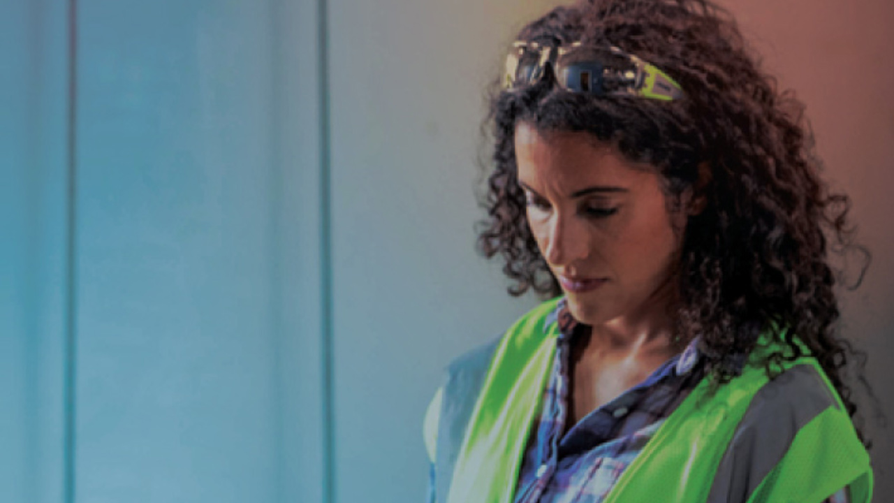 A person with curly hair wearing a neon safety vest and glasses on their head is using a tablet. They are standing against a blue-gray background, appearing focused on the device.