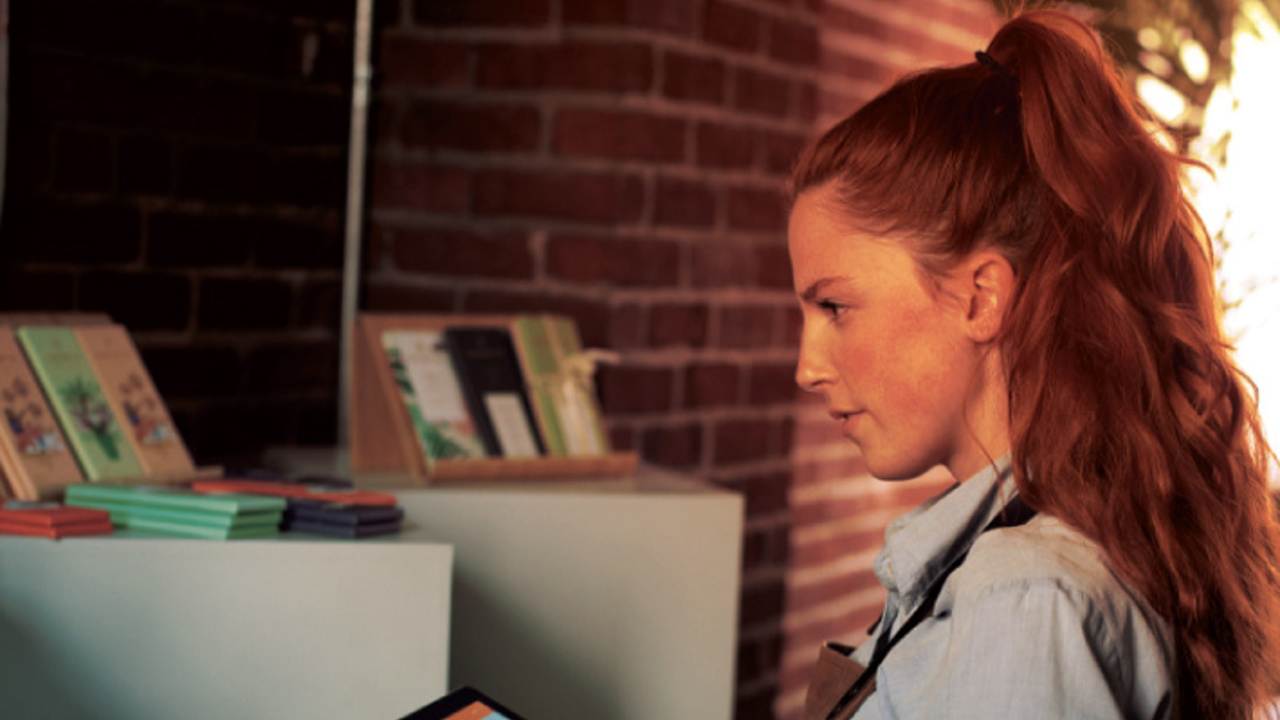 A woman with long red hair, wearing a light blue shirt and apron, holds a tablet while standing in a store. She appears to be checking inventory or product details. Shelves with books or journals are visible in the background.