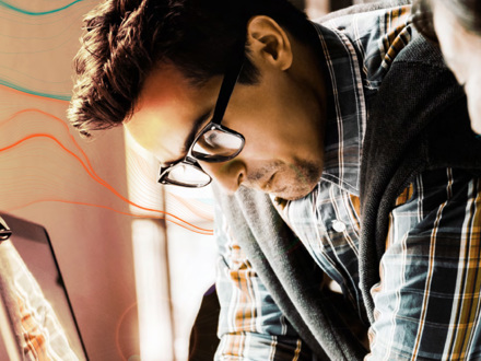 A man with glasses and a plaid shirt, wearing a sweater draped over his shoulders, is focused on something in front of him. Warm lighting and an abstract design with colorful lines create a creative atmosphere.