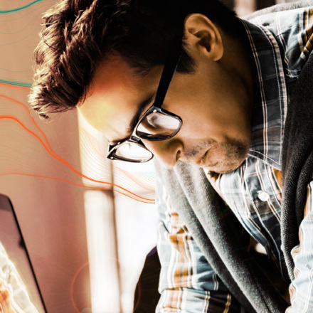 A man with glasses and a plaid shirt, wearing a sweater draped over his shoulders, is focused on something in front of him. Warm lighting and an abstract design with colorful lines create a creative atmosphere.