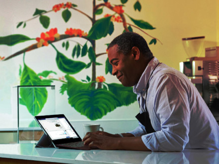 A person in a white shirt and apron is smiling while using a tablet on a countertop. A coffee cup is nearby, and there's a vibrant wall mural behind them depicting coffee plants with leaves and red berries.