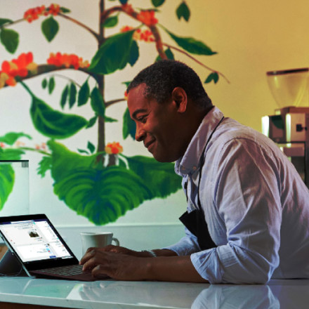 A person in a white shirt and apron is smiling while using a tablet on a countertop. A coffee cup is nearby, and there's a vibrant wall mural behind them depicting coffee plants with leaves and red berries.