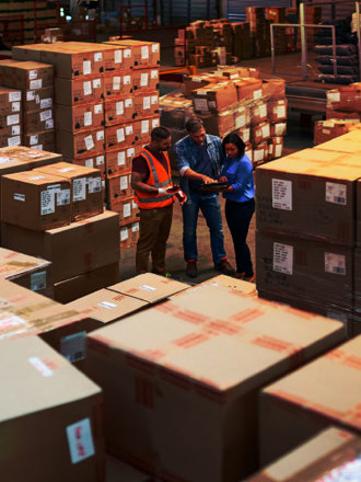 Three warehouse workers stand among stacks of large cardboard boxes. One wears an orange safety vest and helmet, while the others hold a tablet, engaging in a discussion. The warehouse is well-lit, with rows of organized parcels.