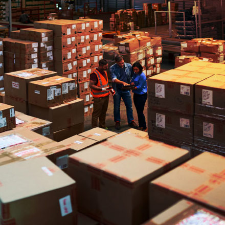 Three warehouse workers stand among stacks of large cardboard boxes. One wears an orange safety vest and helmet, while the others hold a tablet, engaging in a discussion. The warehouse is well-lit, with rows of organized parcels.