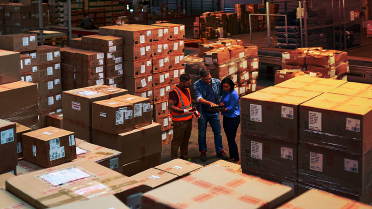 Three warehouse workers stand among stacks of large cardboard boxes. One wears an orange safety vest and helmet, while the others hold a tablet, engaging in a discussion. The warehouse is well-lit, with rows of organized parcels.