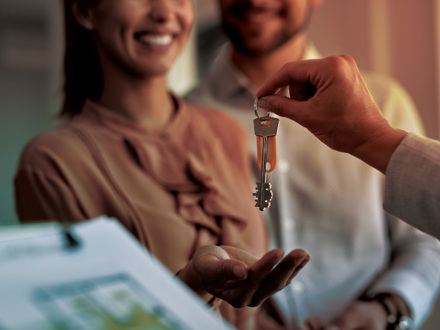 A person hands over a set of keys to a smiling couple. The couple appears happy and excited. In the foreground, there is a clipboard with a document, possibly a contract or agreement.