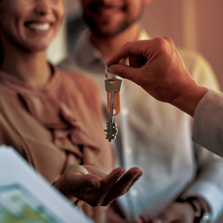 A person hands over a set of keys to a smiling couple. The couple appears happy and excited. In the foreground, there is a clipboard with a document, possibly a contract or agreement.