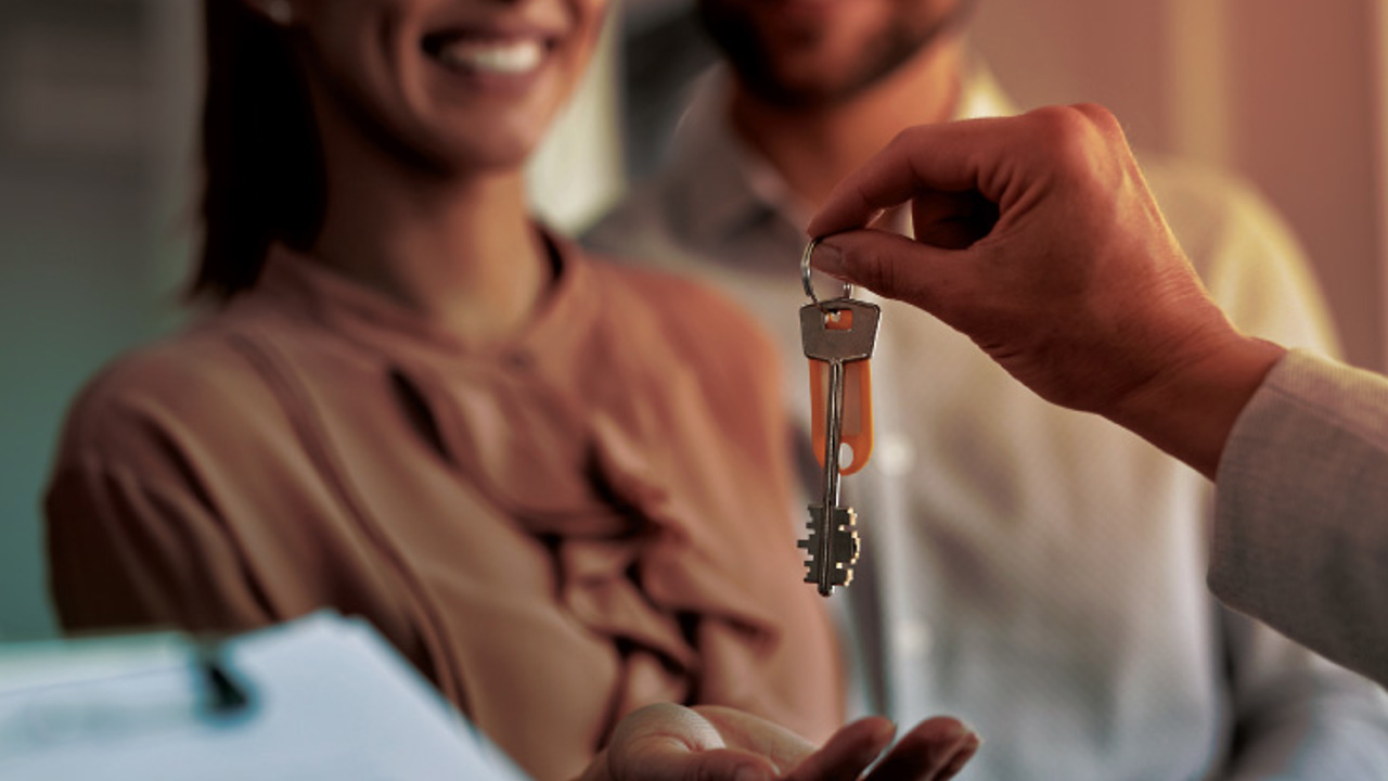 A person hands over a set of keys to a smiling couple. The couple appears happy and excited. In the foreground, there is a clipboard with a document, possibly a contract or agreement.