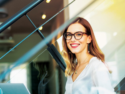 A woman with long hair and glasses smiles while sitting indoors, holding a laptop. She is wearing a white blouse and is by a staircase with modern design elements in the background.