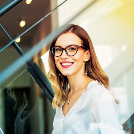 A woman with long hair and glasses smiles while sitting indoors, holding a laptop. She is wearing a white blouse and is by a staircase with modern design elements in the background.