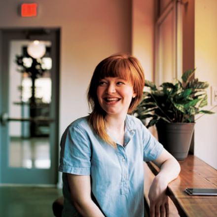 A woman with red hair sits at a wooden table by a window, smiling. She is wearing a light blue shirt. There is a potted plant on the table and a closed door in the background.