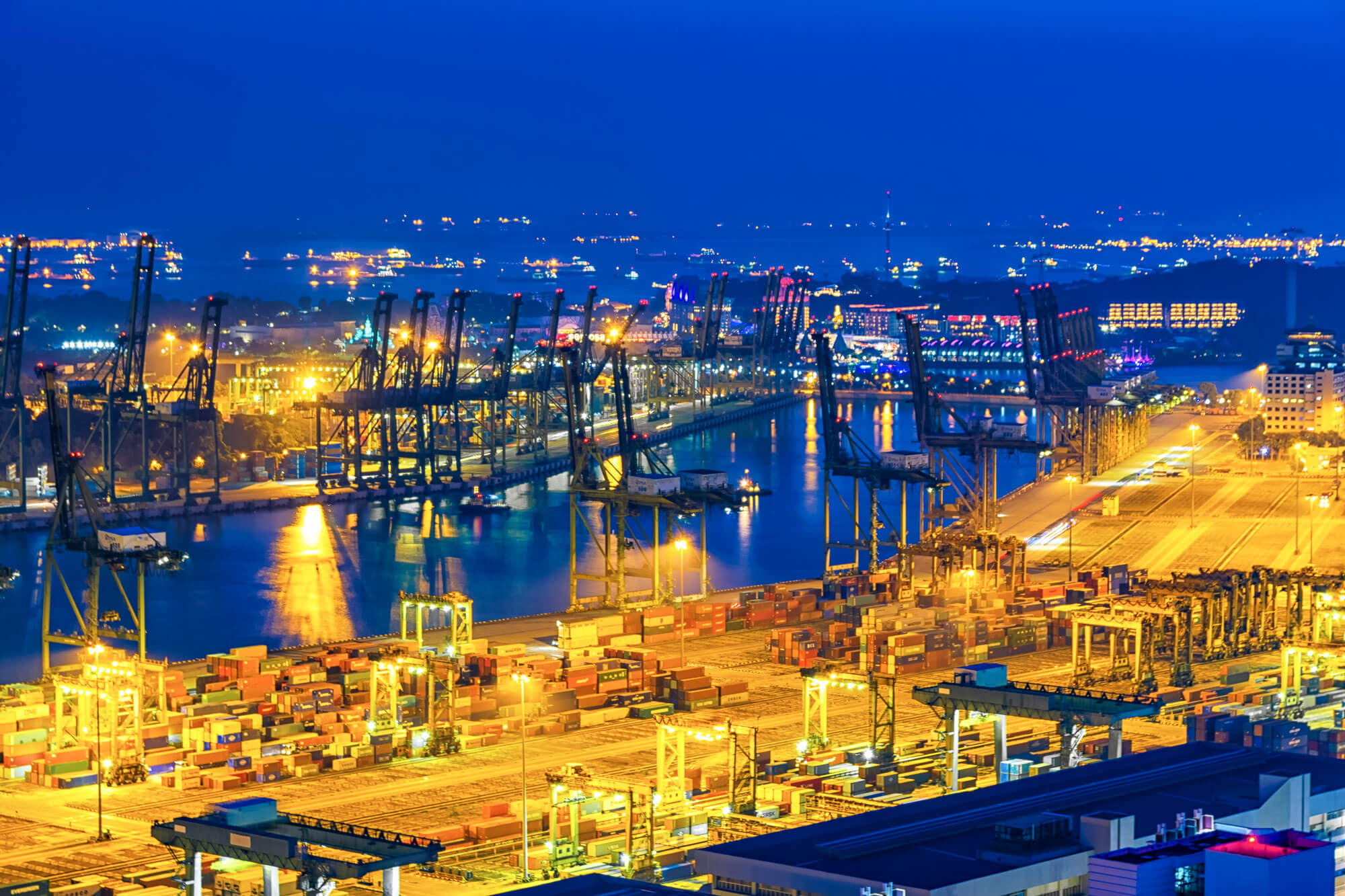 A vibrant, illuminated view of a bustling port at night, with numerous cranes, containers, and docked ships. The bright lights reflect on the water, highlighting the industrial activity against a deep blue sky.