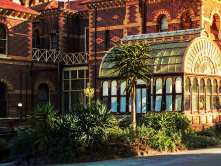 A brick building with ornate designs features a glass conservatory. Lush green plants grow in the garden outside, and the setting sun casts a warm glow on the scene.