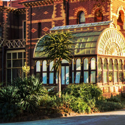 A brick building with ornate designs features a glass conservatory. Lush green plants grow in the garden outside, and the setting sun casts a warm glow on the scene.