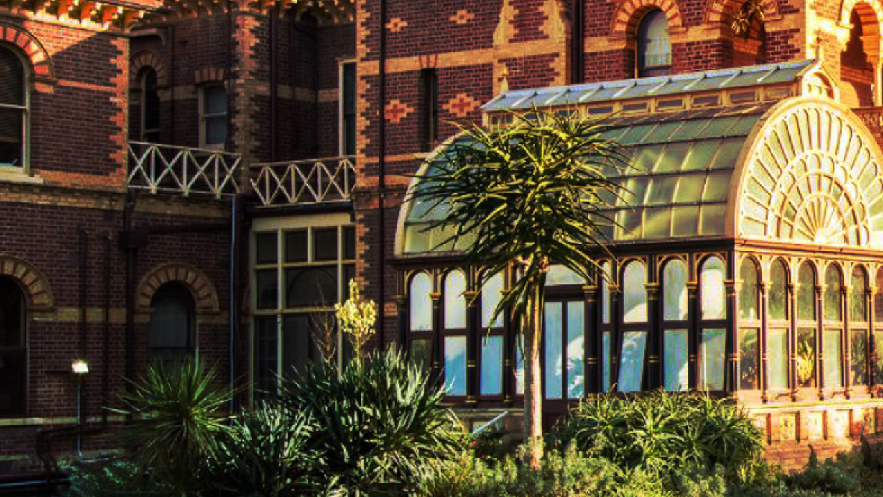 A brick building with ornate designs features a glass conservatory. Lush green plants grow in the garden outside, and the setting sun casts a warm glow on the scene.