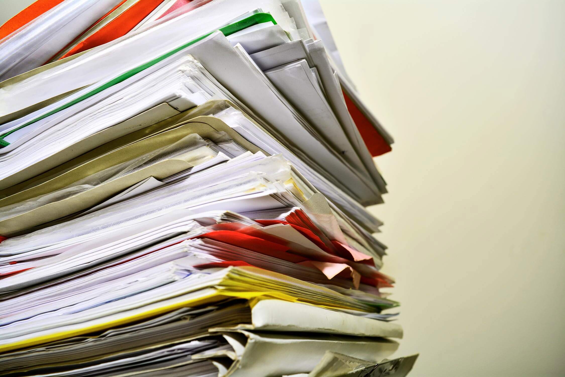 A large stack of papers and folders with various colors, including red, yellow, and green, is piled up against a plain background. Some documents are slightly askew, creating a sense of clutter and urgency.