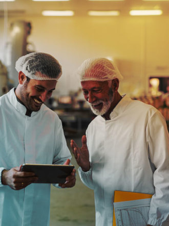 Two cheerful male colleagues in sterile clothes standing in food factory and looking at sales documents on digital tablet.