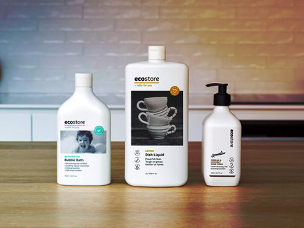 Three bottles of Ecostore products are on a wooden surface. From left to right: Bubble Bath with a child's face, Dish Liquid with stacked teacups, and a hand wash with a pump dispenser. A softly lit kitchen backsplash is in the background.