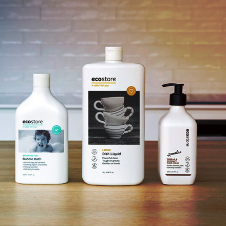 Three bottles of Ecostore products are on a wooden surface. From left to right: Bubble Bath with a child's face, Dish Liquid with stacked teacups, and a hand wash with a pump dispenser. A softly lit kitchen backsplash is in the background.