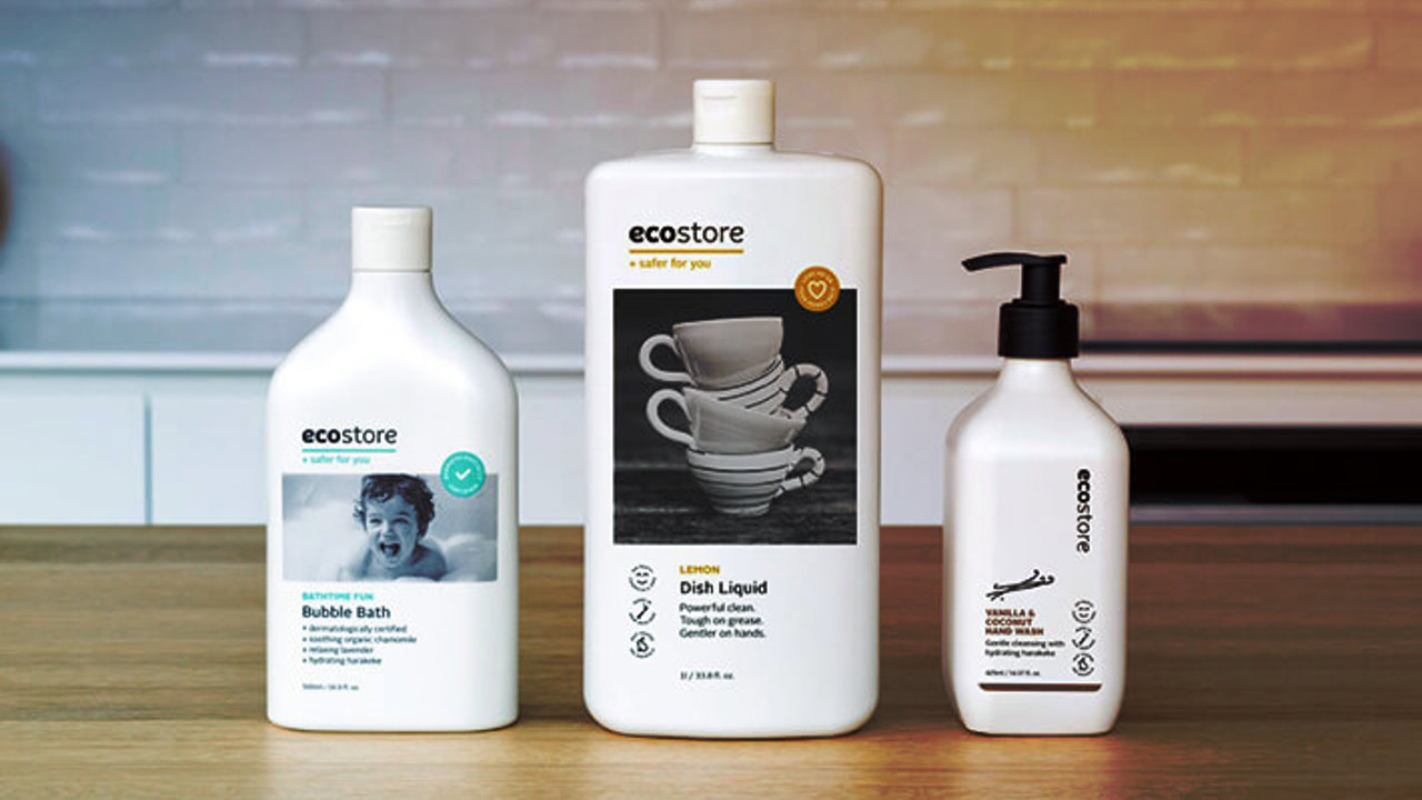 Three bottles of Ecostore products are on a wooden surface. From left to right: Bubble Bath with a child's face, Dish Liquid with stacked teacups, and a hand wash with a pump dispenser. A softly lit kitchen backsplash is in the background.