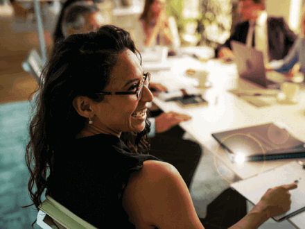 A woman with glasses is smiling during a meeting, seated at a table with notebooks and a laptop. Four other people are blurred in the background, engaged in discussion in a brightly lit office setting.