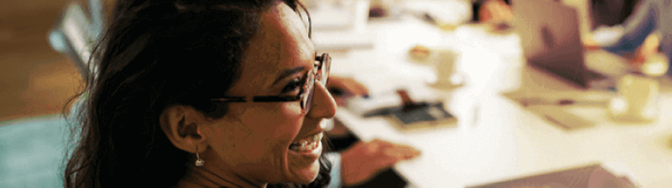 A woman with glasses is smiling during a meeting, seated at a table with notebooks and a laptop. Four other people are blurred in the background, engaged in discussion in a brightly lit office setting.