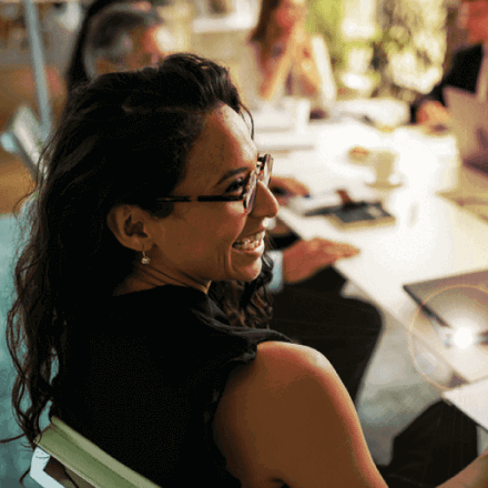 A woman with glasses is smiling during a meeting, seated at a table with notebooks and a laptop. Four other people are blurred in the background, engaged in discussion in a brightly lit office setting.