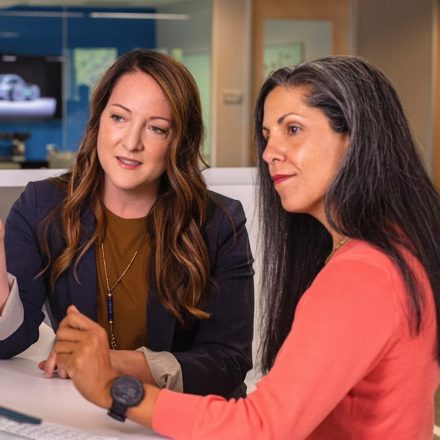 Two Business Women Talking About Sales In Office At Desk With Laptop