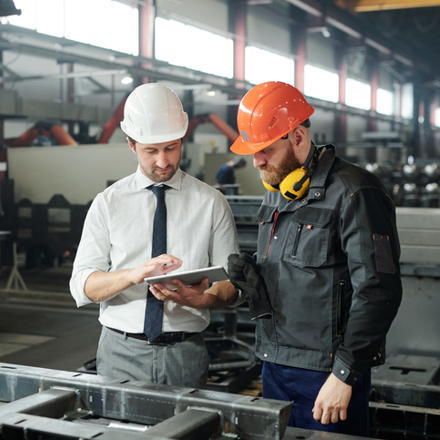 Young master in hardhat and bearded engineer discussing technical sketch on display of tablet in factory workshop.