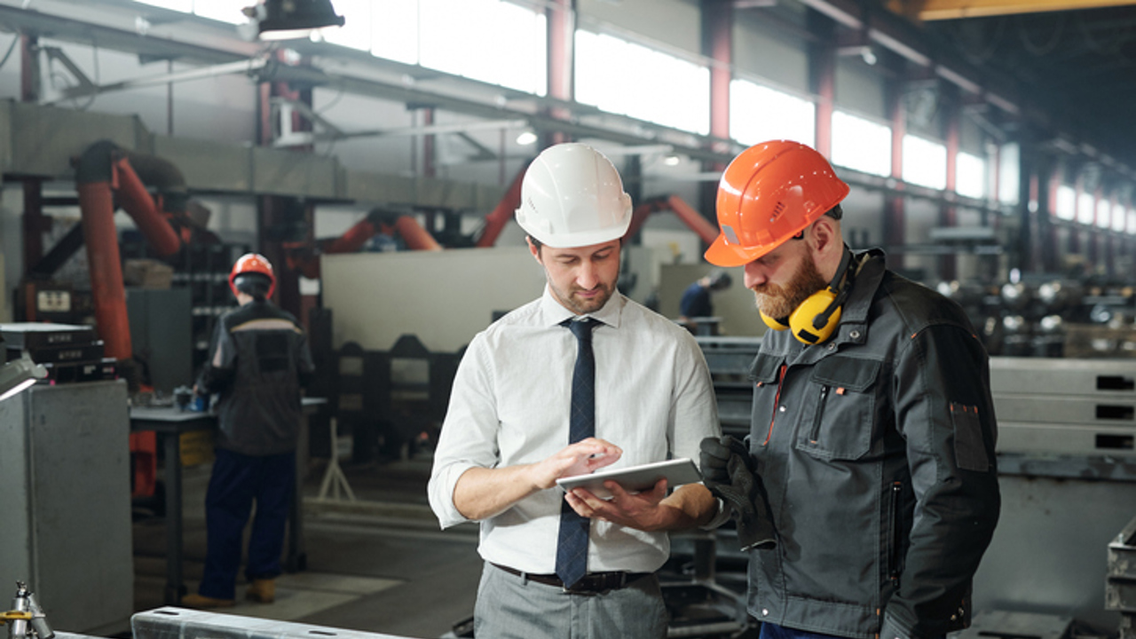 Young master in hardhat and bearded engineer discussing technical sketch on display of tablet in factory workshop.