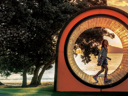 A girl walks through a circular sculpture at sunset, framed by trees and overlooking a tranquil beach with calm water. The orange and transparent structure captures the warm glow of the setting sun.