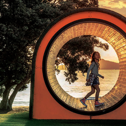 A girl walks through a circular sculpture at sunset, framed by trees and overlooking a tranquil beach with calm water. The orange and transparent structure captures the warm glow of the setting sun.