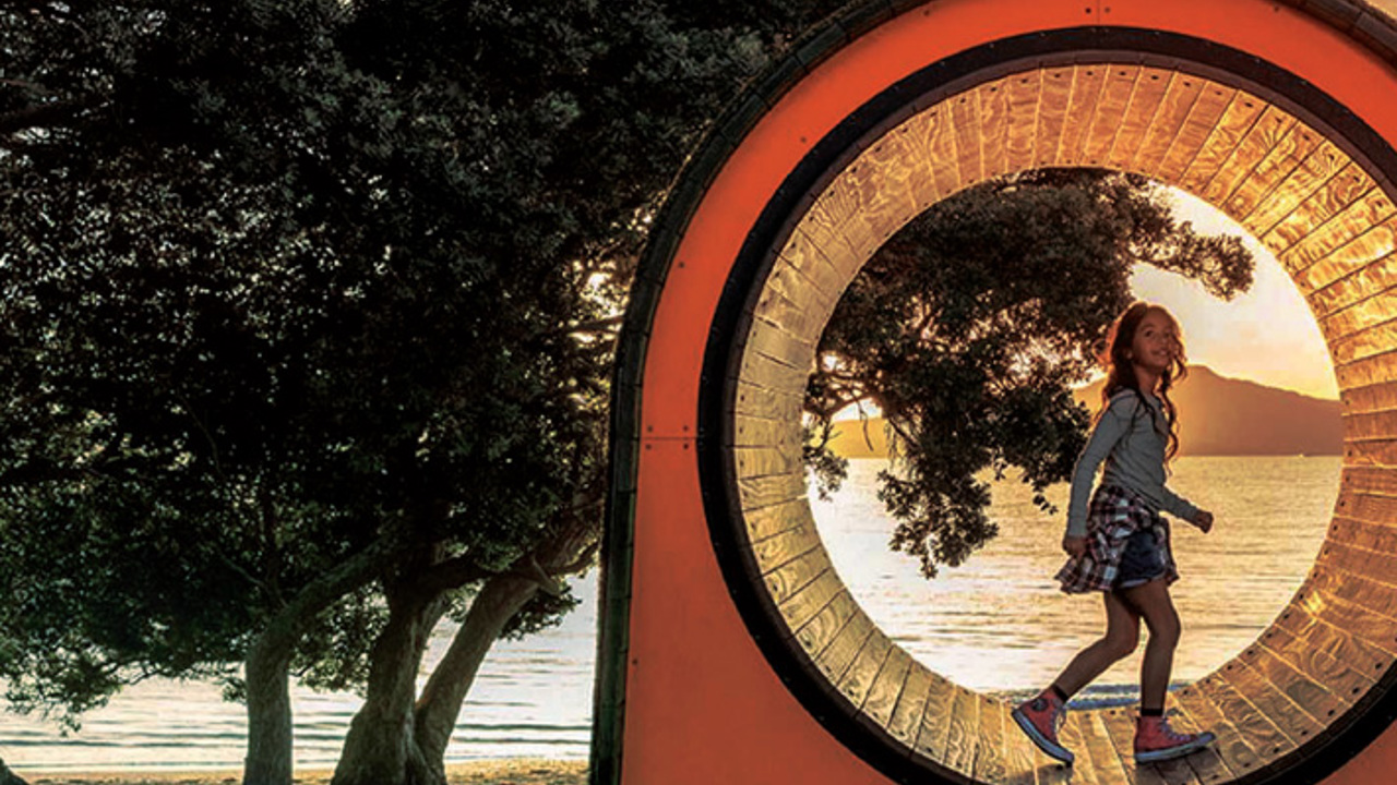 A girl walks through a circular sculpture at sunset, framed by trees and overlooking a tranquil beach with calm water. The orange and transparent structure captures the warm glow of the setting sun.