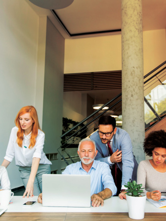 A group of six people sitting and standing around a table in a modern office. They are engaged in discussion, looking at laptops and documents. The room has a staircase and large windows. Small plants are placed on the table, adding a touch of greenery.