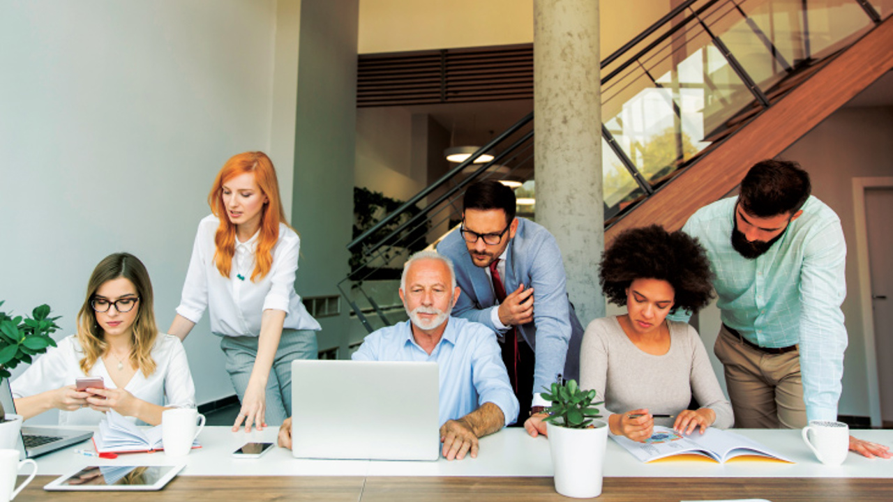 A group of six people sitting and standing around a table in a modern office. They are engaged in discussion, looking at laptops and documents. The room has a staircase and large windows. Small plants are placed on the table, adding a touch of greenery.