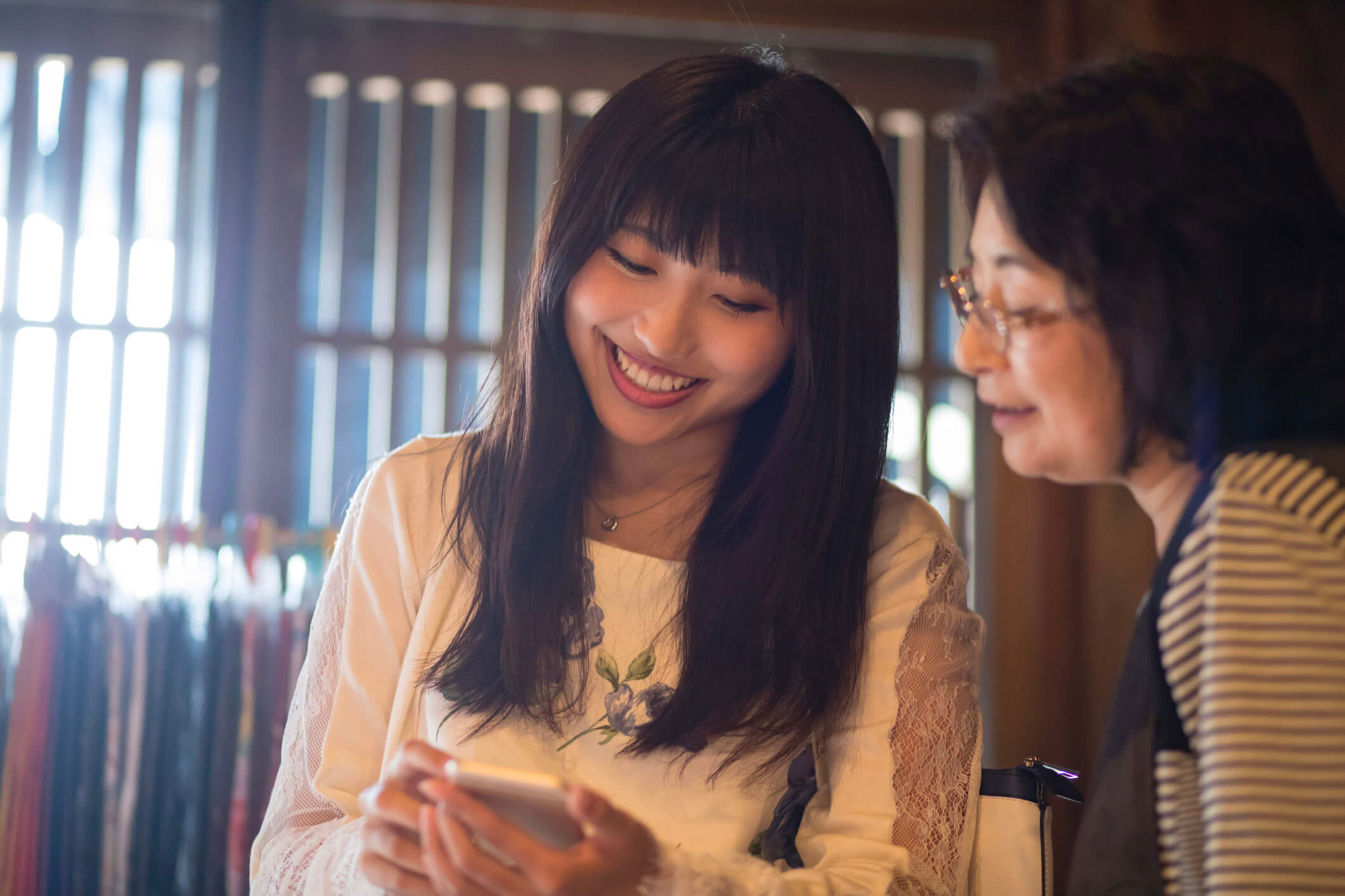 Two women are sitting together, smiling and looking at a smartphone screen. The woman on the left has long dark hair and is wearing a white blouse, while the other woman wears glasses and a striped top. The background has wooden elements and blurred clothing.