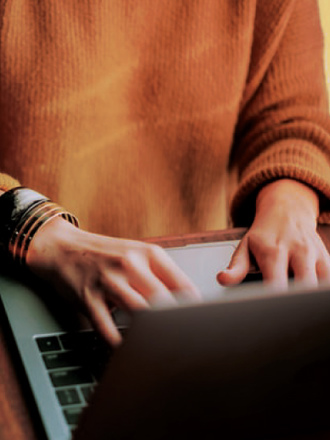 A person wearing a brown sweater types on a laptop at a wooden desk. They are wearing several bracelets on their wrist.