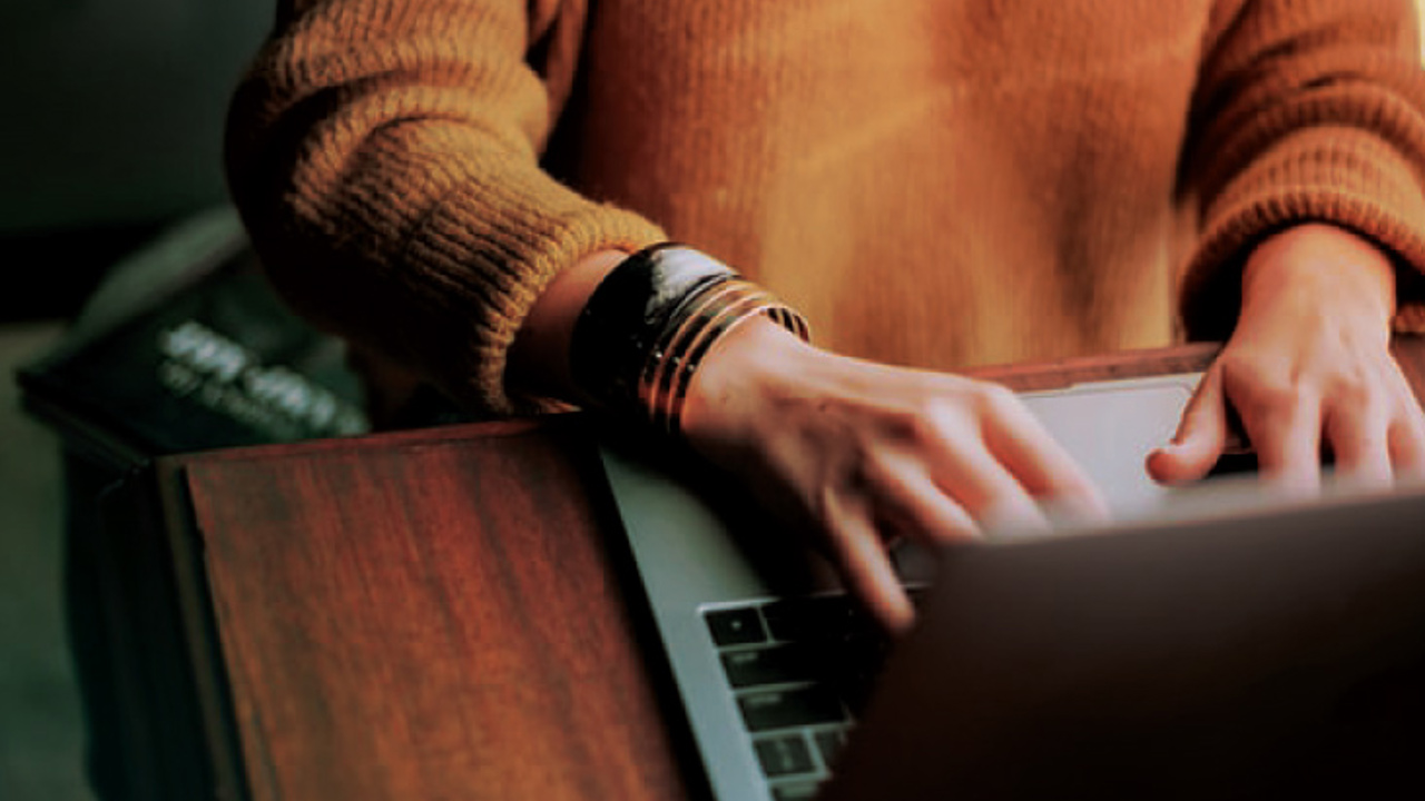 A person wearing a brown sweater types on a laptop at a wooden desk. They are wearing several bracelets on their wrist.