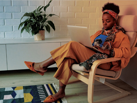 A person sits on a chair, using a laptop while dressed in orange clothing and a colorful scarf. A potted plant is on a cabinet behind them. A patterned rug is on the wooden floor.