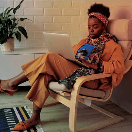 A person sits on a chair, using a laptop while dressed in orange clothing and a colorful scarf. A potted plant is on a cabinet behind them. A patterned rug is on the wooden floor.