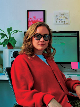 A person with shoulder-length hair and glasses sits confidently in an office chair, wearing a red cardigan. They are in front of dual monitors displaying code and a web page, with plants and colorful sticky notes on the desk.