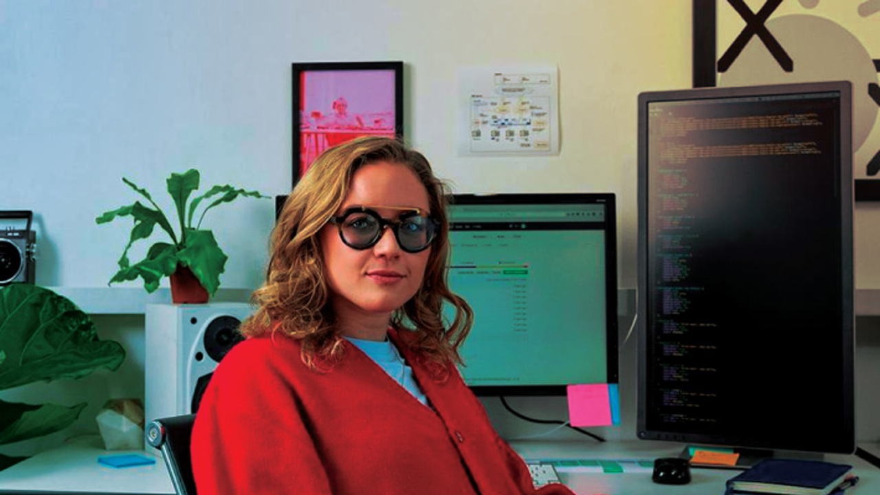 A person with shoulder-length hair and glasses sits confidently in an office chair, wearing a red cardigan. They are in front of dual monitors displaying code and a web page, with plants and colorful sticky notes on the desk.