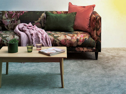 A floral-patterned sofa with green and pink cushions sits against a wall. A pink blanket is draped over the arm. In front, a wooden coffee table holds two glasses and a magazine. A small potted plant is on the table, and the carpet is light gray.