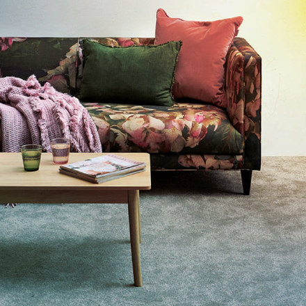 A floral-patterned sofa with green and pink cushions sits against a wall. A pink blanket is draped over the arm. In front, a wooden coffee table holds two glasses and a magazine. A small potted plant is on the table, and the carpet is light gray.