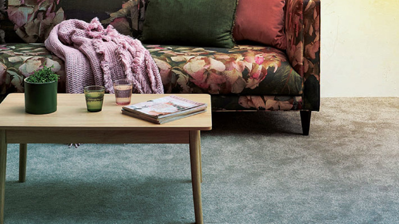 A floral-patterned sofa with green and pink cushions sits against a wall. A pink blanket is draped over the arm. In front, a wooden coffee table holds two glasses and a magazine. A small potted plant is on the table, and the carpet is light gray.