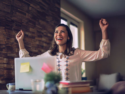 A woman in a white blouse is sitting at a desk with a laptop, looking joyful and raising her arms in celebration. The laptop has colorful sticky notes on it, and the background is softly lit.