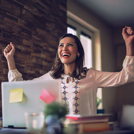 A woman in a white blouse is sitting at a desk with a laptop, looking joyful and raising her arms in celebration. The laptop has colorful sticky notes on it, and the background is softly lit.