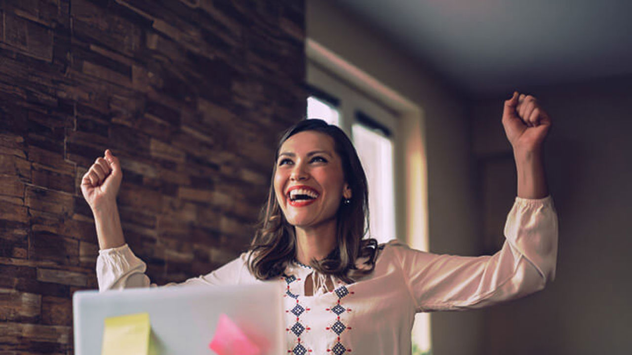 A woman in a white blouse is sitting at a desk with a laptop, looking joyful and raising her arms in celebration. The laptop has colorful sticky notes on it, and the background is softly lit.